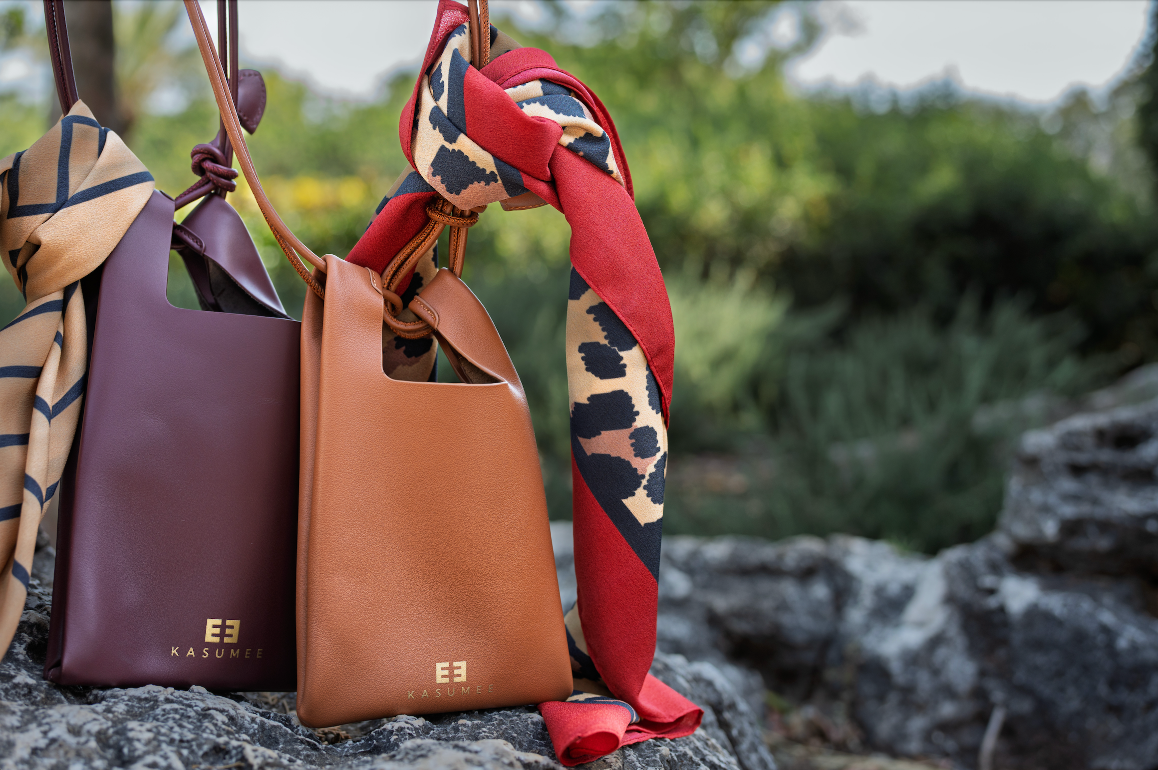 Two luxury Kasumee leather crossbody phone bags in burgundy and caramel, each styled with a patterned scarf, displayed on textured stone with a blurred natural background.