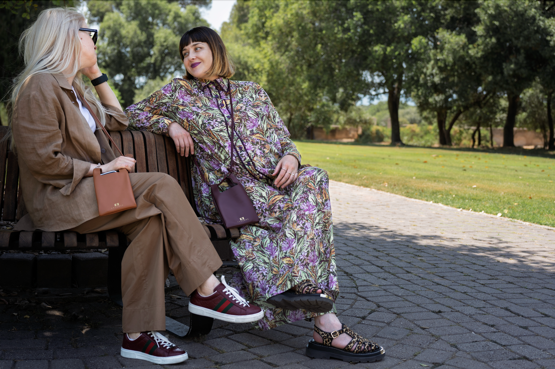An elegant park scene featuring two fashionable women in chic outfits, effortlessly showcasing luxury Kasumee leather crossbody bags in burgundy and caramel colors against a backdrop of sunlit trees and lush greenery.