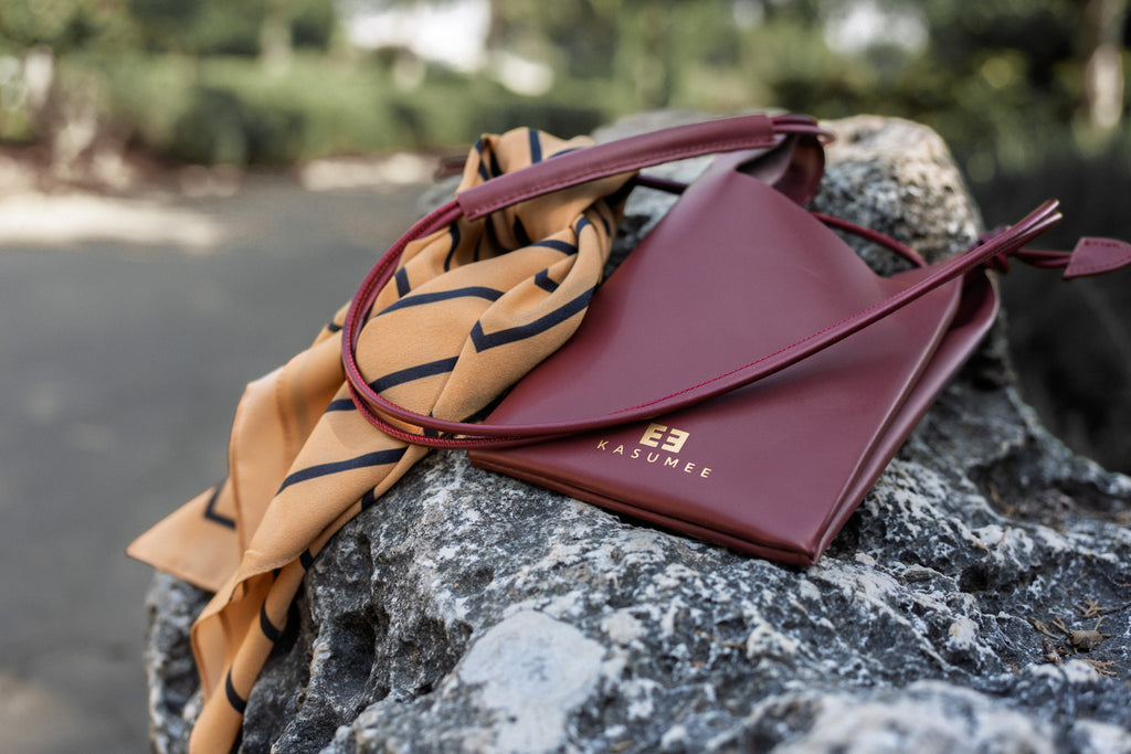 A luxury Kasumee burgundy leather crossbody phone bag with gold logo detail, elegantly styled with a mustard and black striped scarf, resting on a textured stone surface in an outdoor setting.