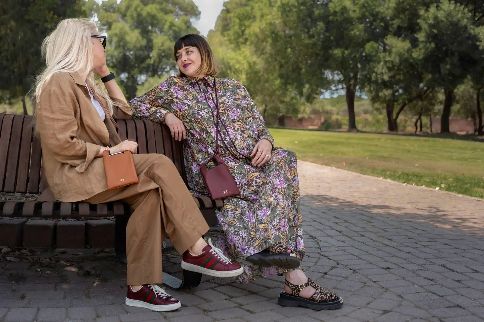 Two women sitting on a park bench, wearing stylish fashion outfits. One is in a tan blazer and trousers with red sneakers and a brown handbag, while the other is in a floral maxi dress with red sneakers and a brown crossbody bag.